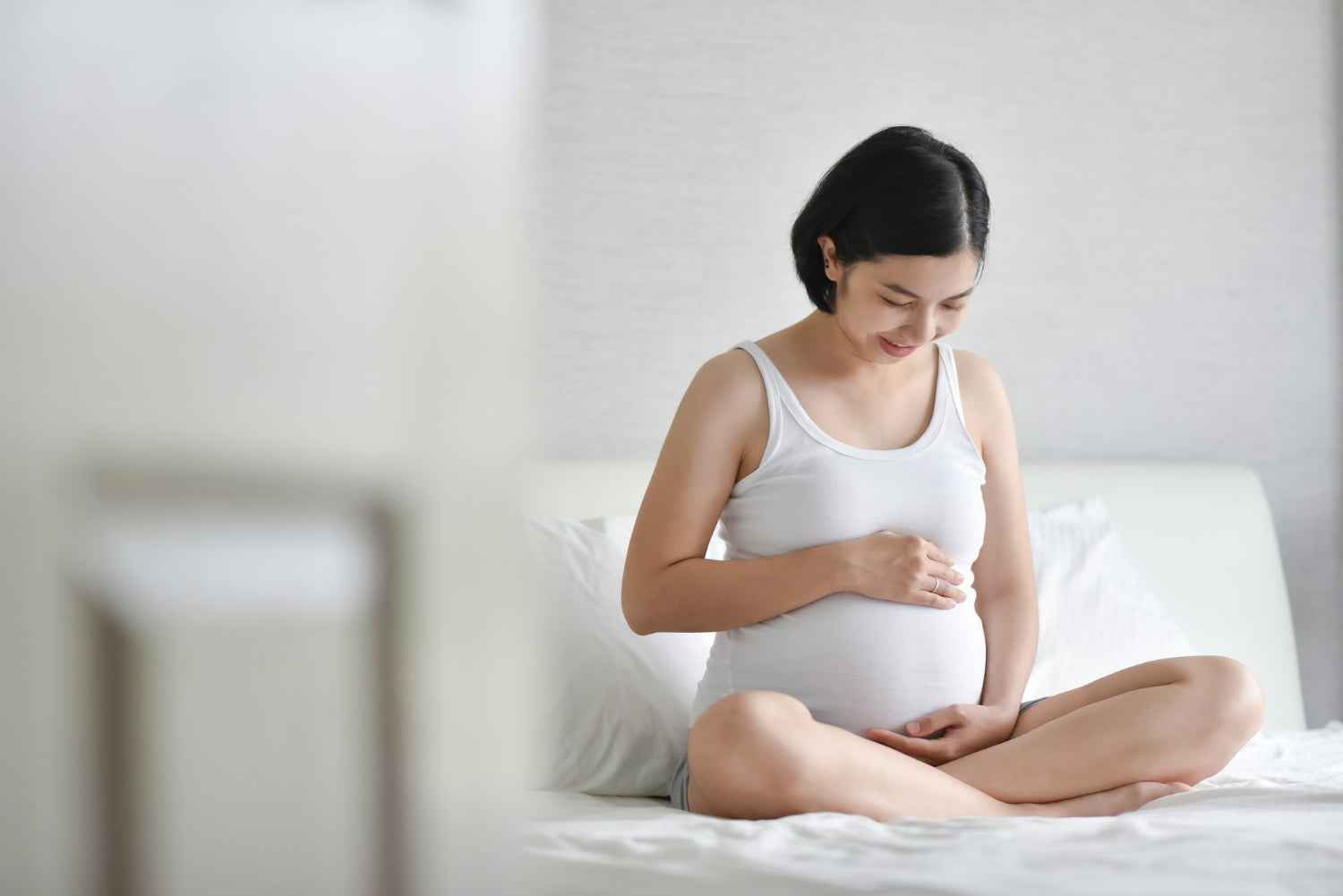 Pregnant woman sitting on a bed in a white tank top, looking at her belly.